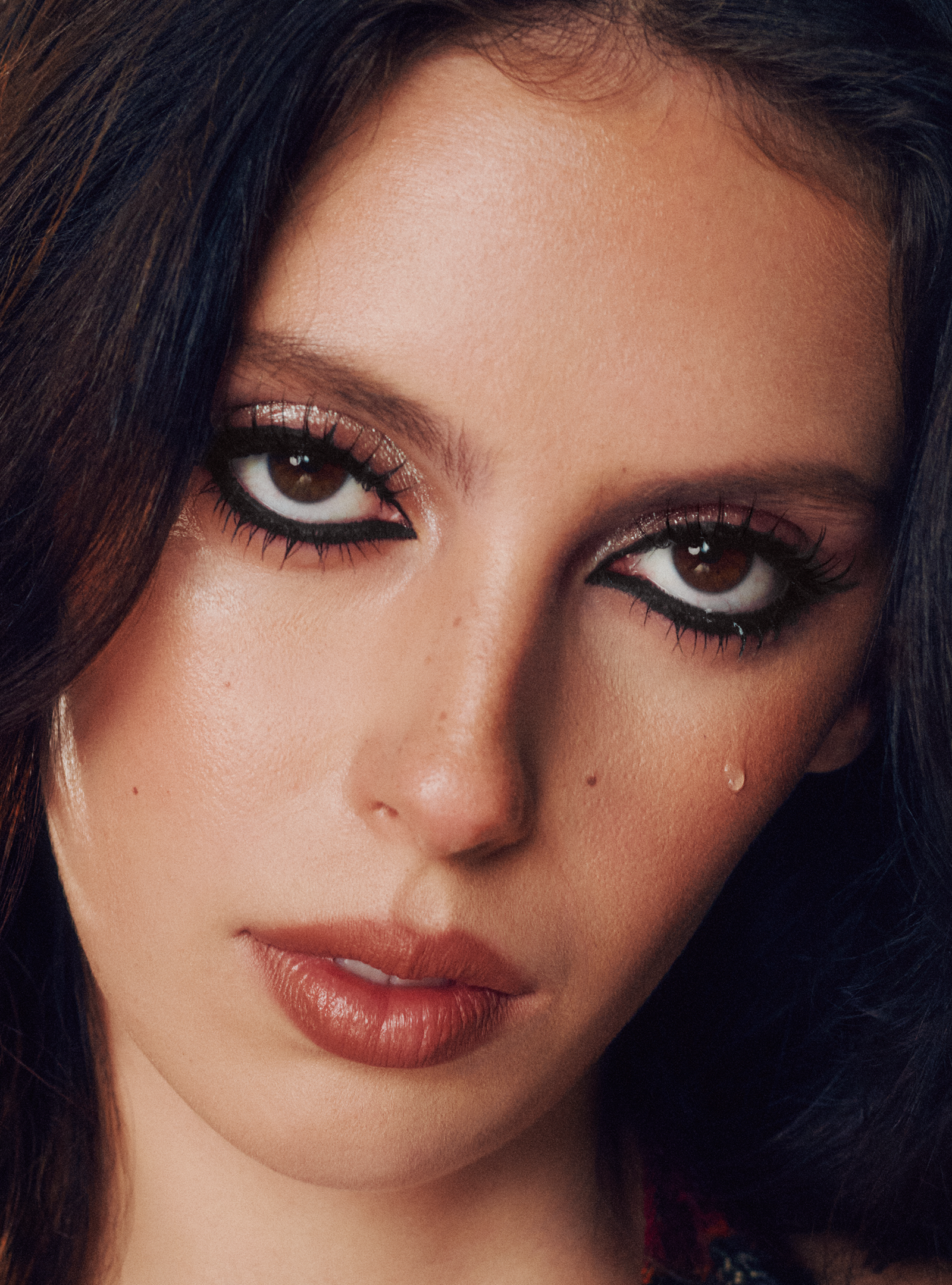 Close-up portrait of a woman with dramatic black eye makeup, long dark hair, and nude lips against a dark background.